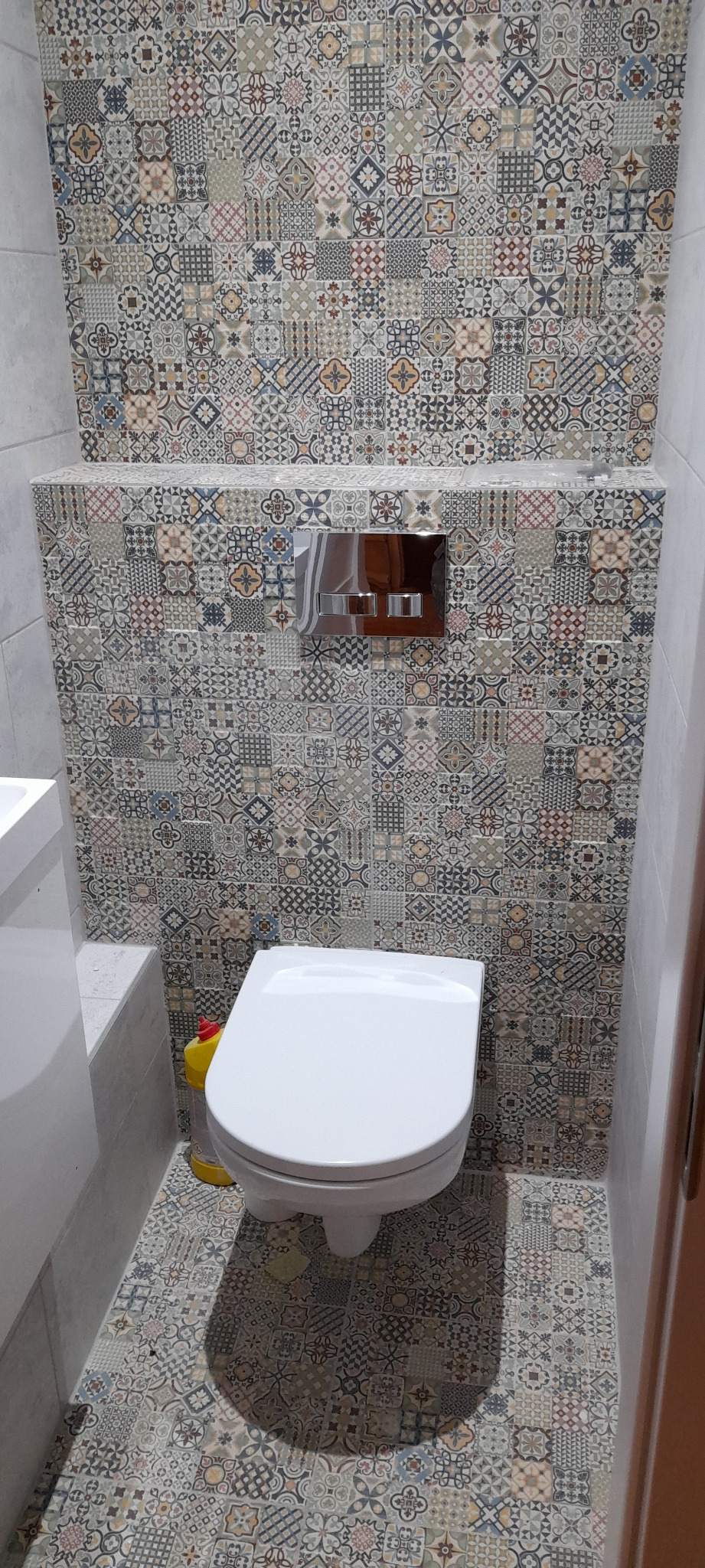 Nowoczesna toaleta podwieszana z chromowanym przyciskiem spłukującym, otoczona ścianami i podłogą wyłożonymi dekoracyjnymi płytkami w stylu patchwork.