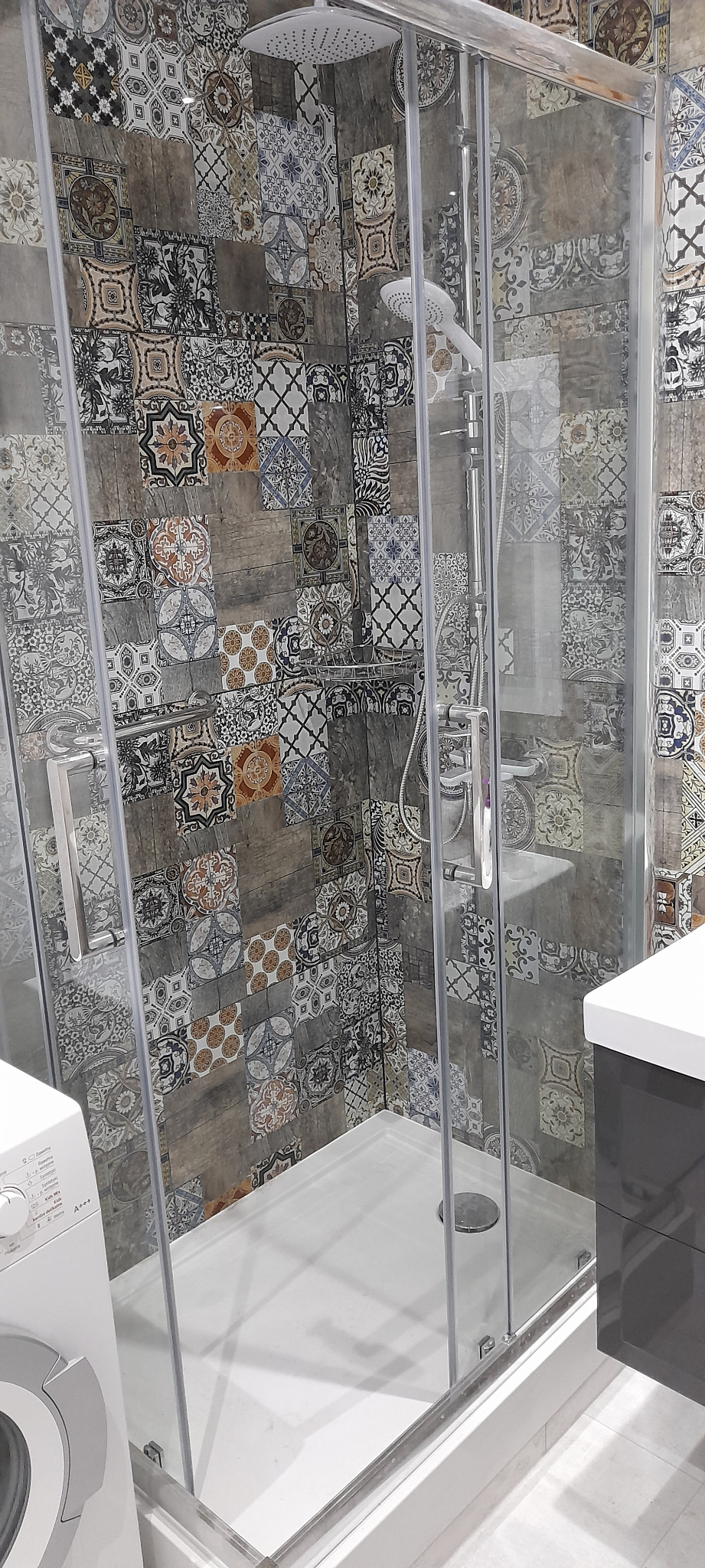 Nowoczesna kabina prysznicowa z deszczownicą, obudowana płytkami w stylu patchwork, widoczna pralka i umywalka z szafką w tle.
