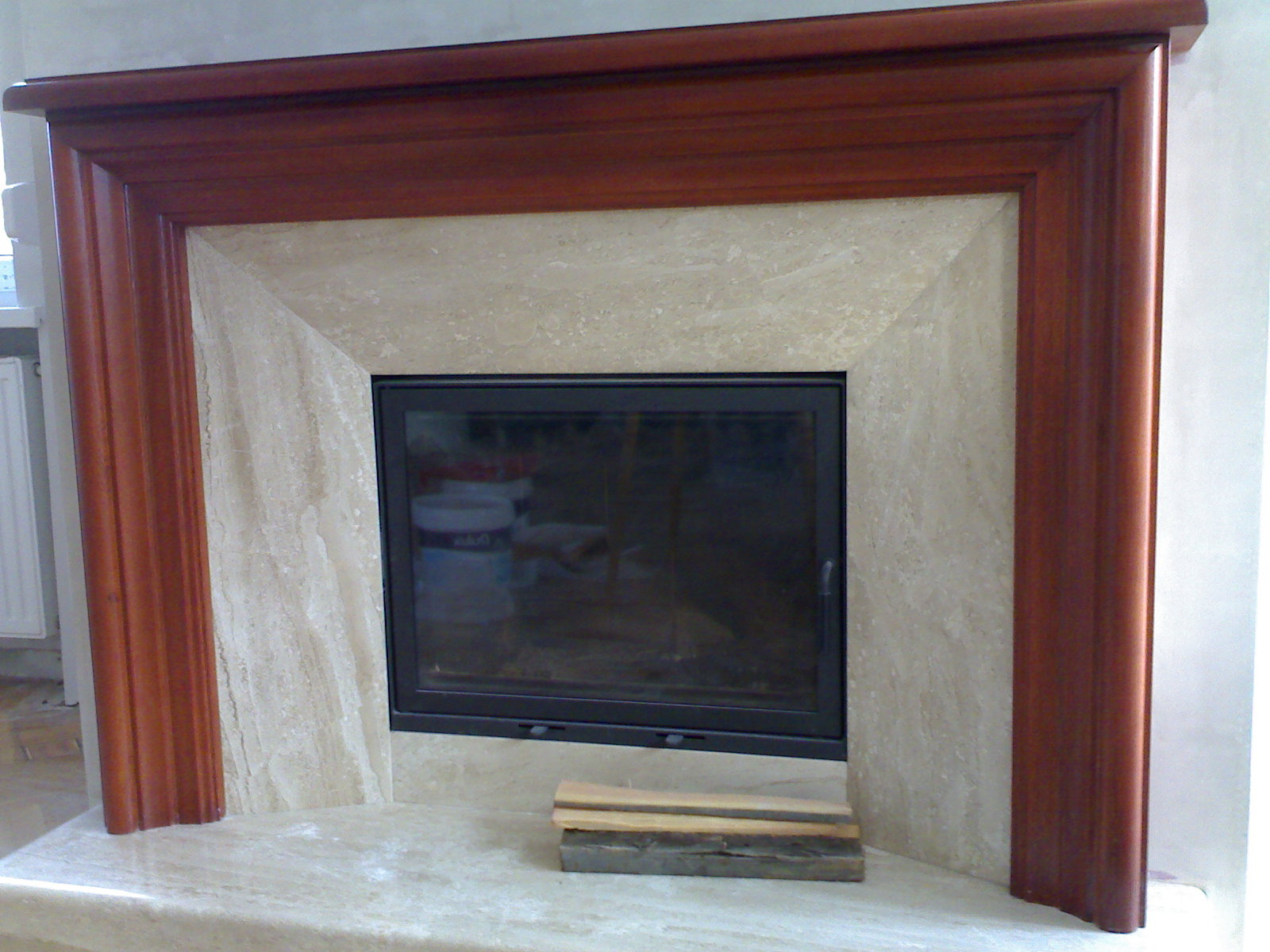 Obudowa kominka z marmuru i drewna, z widocznym wkładem kominkowym i ułożonymi obok szczapami drewna.