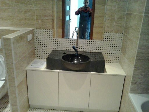 Nowoczesna łazienka z umywalką w kształcie misy na blacie z ciemnego kamienia, białe szafki pod umywalką, beżowe płytki na ścianach i lustro, w którym odbija się osoba robiąca zdjęcie.