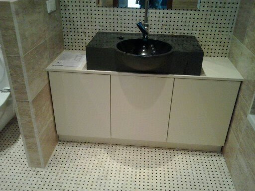 Wykończona łazienka z czarną, okrągłą umywalką nablatową na szafce z trzema białymi frontami, jasne płytki na ścianach i podłodze z drobnym wzorem.