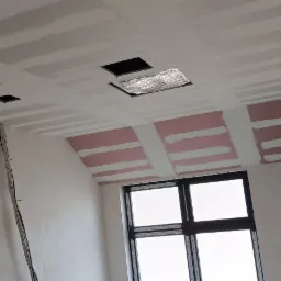 Montaż sufitu podwieszanego z widocznymi profilami i otworami na oświetlenie, częściowo wykończone ściany w surowym stanie, okno w tle.