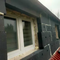 Ocieplanie budynku styropianem grafitowym, widoczne białe okno PCV, piana montażowa uszczelniająca oraz czerwona blacha dachowa