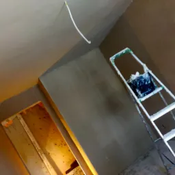 Wnętrze w trakcie prac wykończeniowych: świeżo nałożona szpachla na ścianach i suficie, drabina malarska z wiadrem w rogu pokoju, wystający kabel elektryczny.