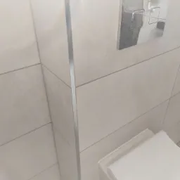 Białe, poziome płytki w łazience z chromowaną listwą narożną i przyciskiem spłuczki podtynkowym z widoczną instalacją hydrauliczną.