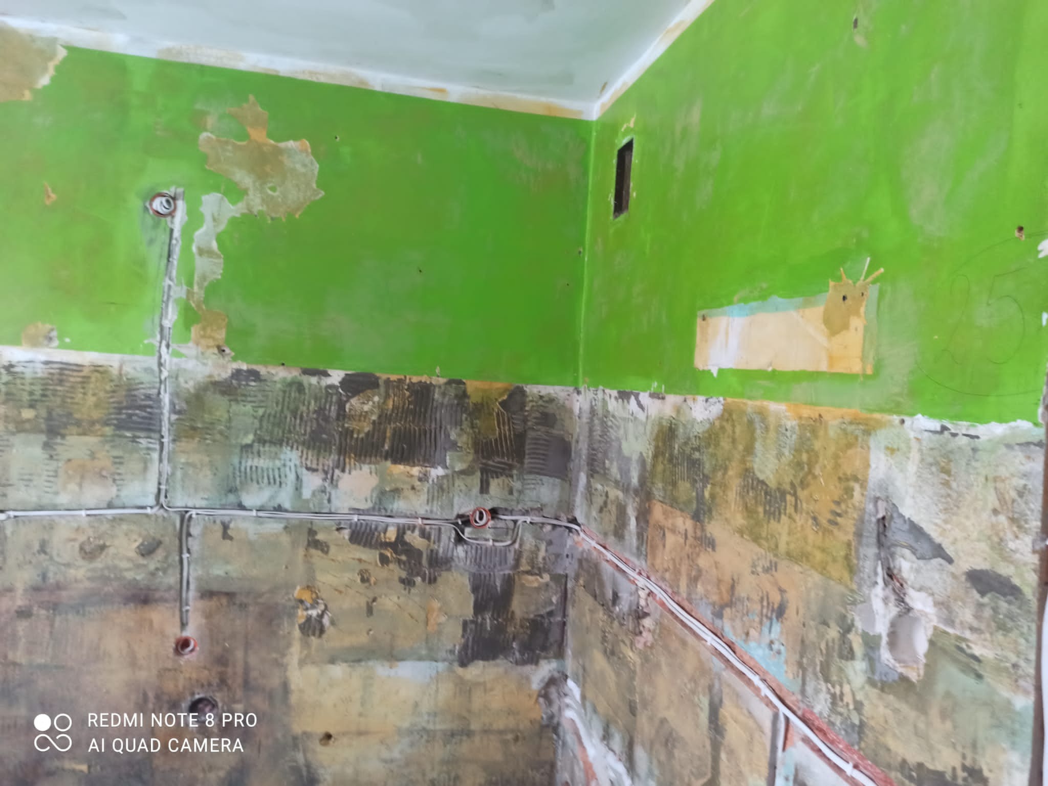 Ściana w trakcie remontu z widocznymi przewodami elektrycznymi, fragmentami zielonej farby i resztkami starej okładziny, przygotowana do dalszych prac wykończeniowych.