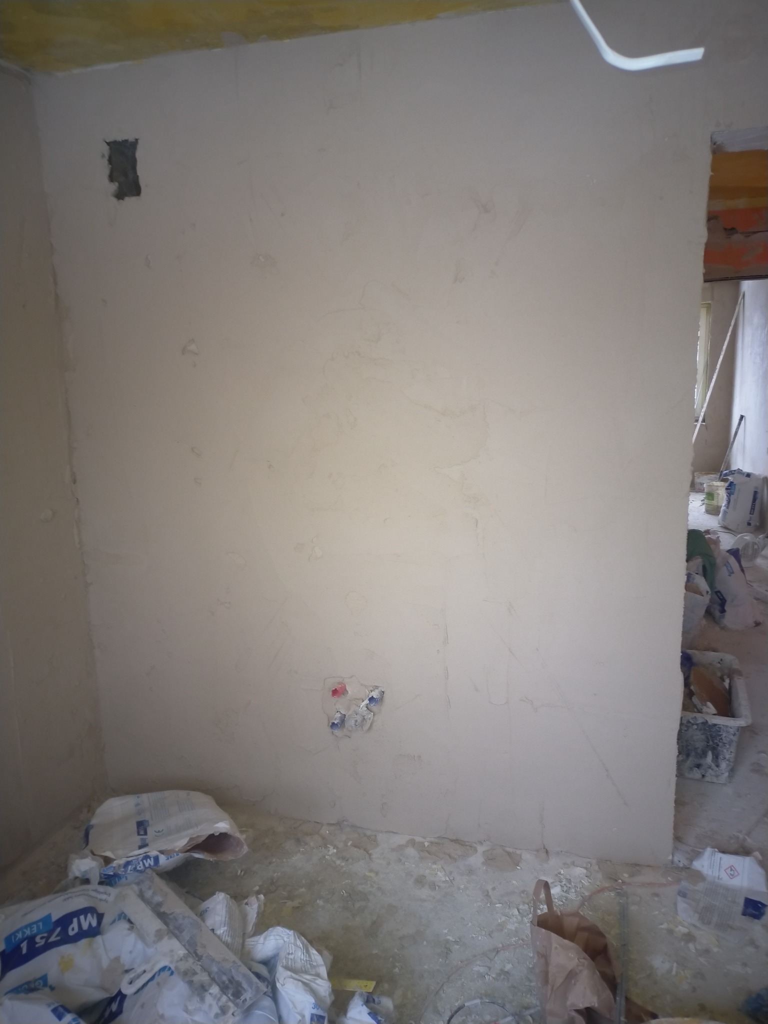 Ściana po wstępnym szpachlowaniu z widocznymi niedoskonałościami, przygotowana do dalszych prac wykończeniowych; w tle worki z zaprawą gipsową i inne materiały budowlane.