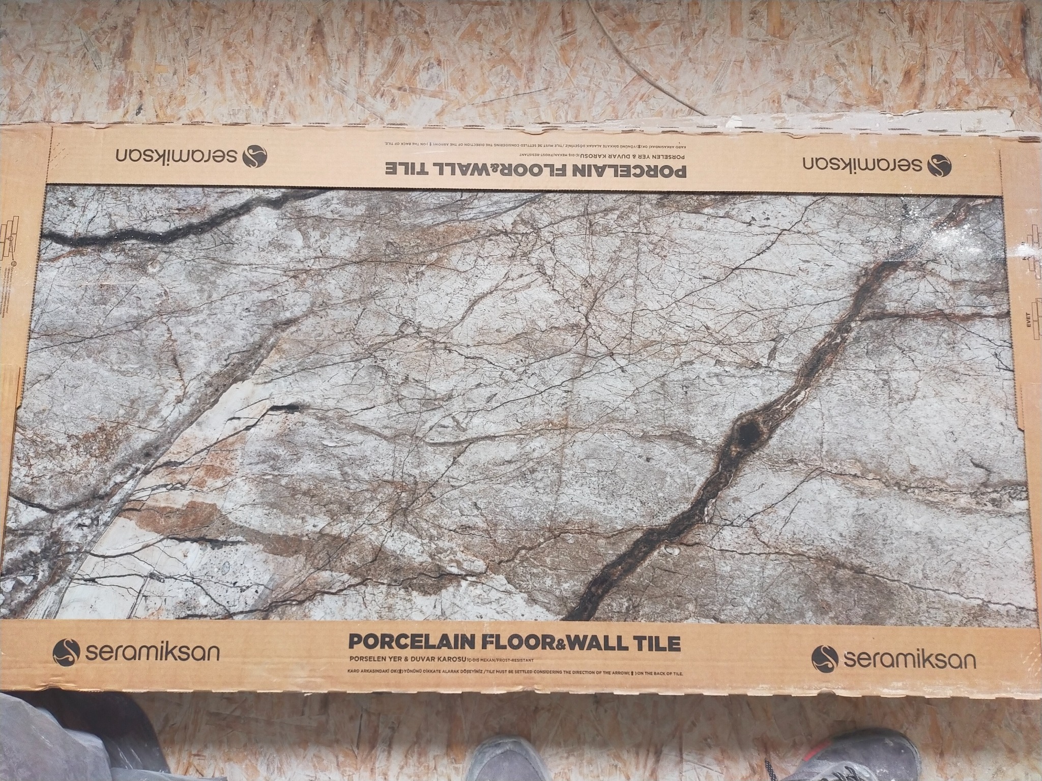 Płytka gresowa podłogowo-ścienna Seramiksan w kartonie, wzór imitujący marmur z wyraźnym, ciemnym żyłkowaniem, widoczne fragmenty butów osoby fotografującej.
