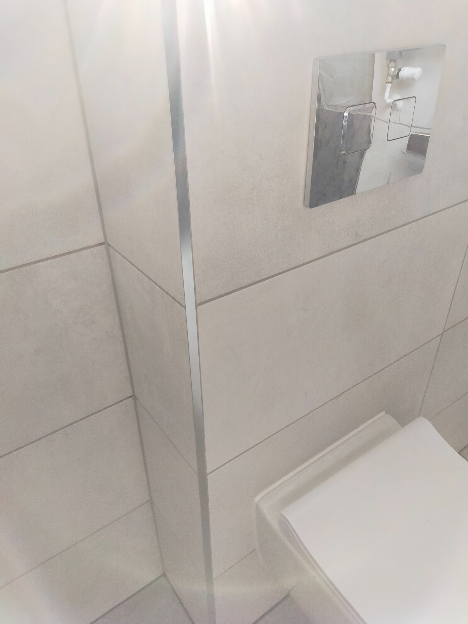 Białe, poziome płytki w łazience z chromowaną listwą narożną i przyciskiem spłuczki podtynkowym z widoczną instalacją hydrauliczną.