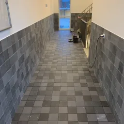 Korytarz wyłożony szarą kwadratową glazurą na podłodze i ścianach do połowy wysokości; widoczne narzędzia glazurnicze, gniazdko elektryczne na ścianie i okno z widokiem na budynek.