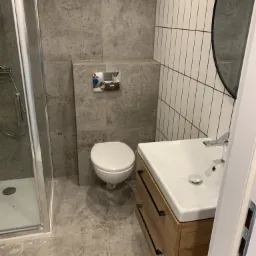 Wykończona łazienka w Pile z kabiną prysznicową, toaletą i umywalką z drewnianą szafką. Ściany wyłożone płytkami imitującymi beton i białymi, wąskimi płytkami z czarną fugą.