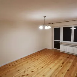 Jasne, puste wnętrze po remoncie: drewniana podłoga, świeżo pomalowane ściany w odcieniach beżu, biały grzejnik pod oknem i ozdobna lampa sufitowa.