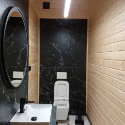 Wąska toaleta z drewnianymi panelami, czarnym marmurem na ścianie i podłużnym oświetleniem, widok na umywalkę z czarną armaturą, lustro i toaletę.