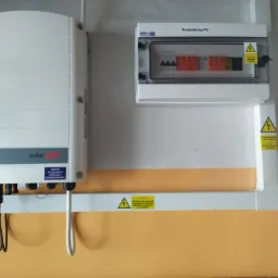 Biały inwerter fotowoltaiczny SolarEdge z podłączonymi kablami oraz rozdzielnica PV na ścianie o pomarańczowym pasie, widoczne naklejki ostrzegawcze o wysokim napięciu.