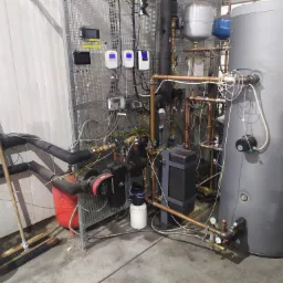 Kotłownia z szarym zbiornikiem akumulacyjnym, pompą obiegową Grundfos, orurowaniem miedzianym i grzejnikami, widoczny panel sterowania na perforowanej ścianie oraz miotła w rogu pomieszczenia.