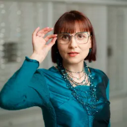 Kobieta z rudymi włosami w turkusowej bluzce i biżuterii, przymierza metalowe okulary na tle ściany z ekspozycją oprawek.