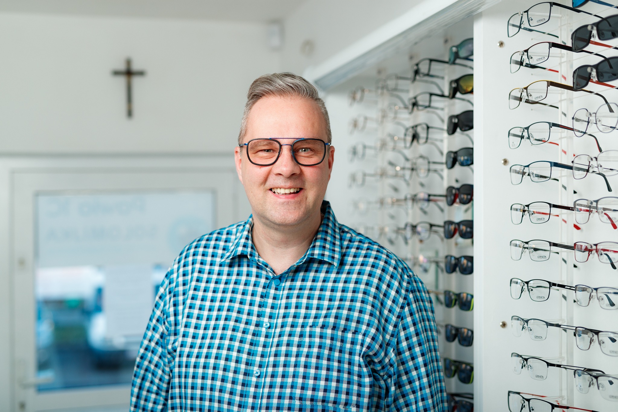 Uśmiechnięty mężczyzna w okularach na tle ekspozycji oprawek okularowych w salonie optycznym, widoczny krzyż na ścianie.