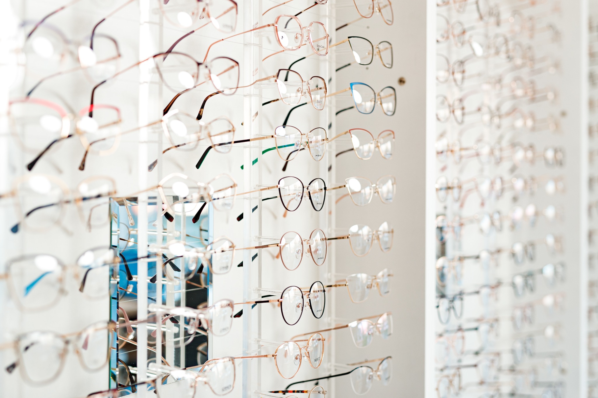 Różnorodne oprawki okularowe na ekspozytorze w salonie optycznym, dominują cienkie, metalowe ramki w odcieniach złota i brązu, niektóre z kolorowymi akcentami.
