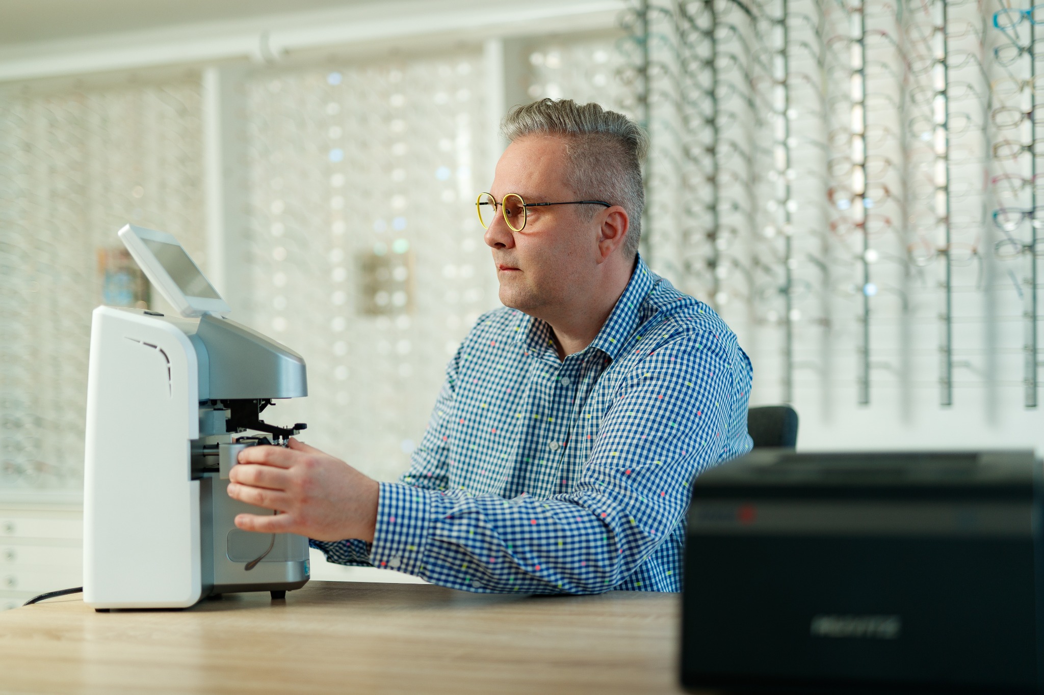 Mężczyzna w okularach o żółtych oprawkach obsługuje autorefraktometr w gabinecie optycznym, w tle ściana z ekspozycją oprawek.
