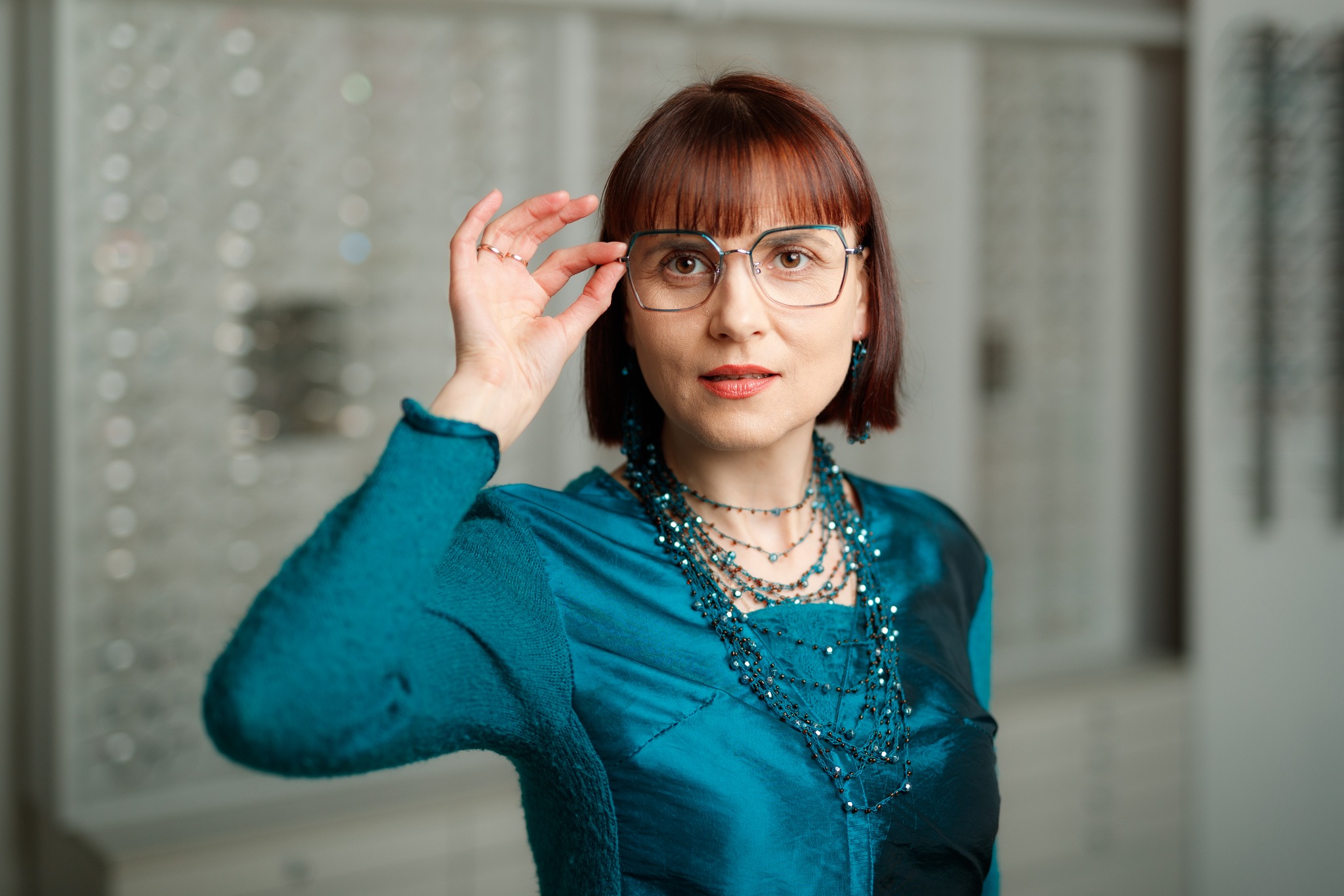 Kobieta z rudymi włosami w turkusowej bluzce i biżuterii, przymierza metalowe okulary na tle ściany z ekspozycją oprawek.