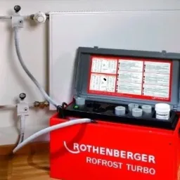 Zamrażarka rur Rothenberger ROFROST TURBO podłączona do grzejnika, widoczne przewody i zawory termostatyczne w białej obudowie.