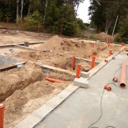 LUK-BUD Łukasz Hachuła - Fundamenty domu w trakcie budowy z widocznymi pomarańczowymi rurami kanalizacyjnymi ułożonymi w wykopach, pracownik łączy rury, w tle drzewa.