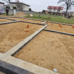 Przygotowanie fundamentów pod budynek z widocznymi pionami kanalizacyjnymi i izolacją przeciwwilgociową, z samochodem firmy hydraulicznej w tle.