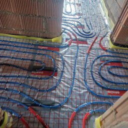 LUK-BUD Łukasz Hachuła - Układanie niebieskich i czerwonych rur ogrzewania podłogowego na folii z siatką, widoczne fragmenty muru z cegieł.