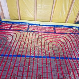 LUK-BUD Łukasz Hachuła - Czerwone rury ogrzewania podłogowego, mocowane zielonymi uchwytami do folii z nadrukowaną siatką, ułożone na żółtej izolacji przy ścianie z drewnianą konstrukcją.