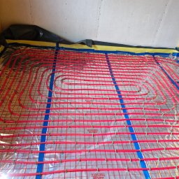 LUK-BUD Łukasz Hachuła - Czerwone rury ogrzewania podłogowego ułożone na folii refleksyjnej z siatką, przymocowane zielonymi klipsami i niebieską taśmą do podłoża, przygotowane do zalania wylewką.