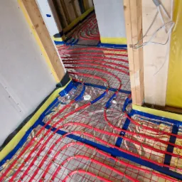 Czerwone rury ogrzewania podłogowego przymocowane do siatki na folii izolacyjnej, widoczne elementy konstrukcji budynku, żółta pianka izolacyjna przy ścianach.