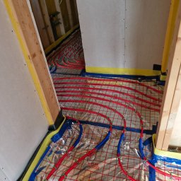 LUK-BUD Łukasz Hachuła - Czerwone i niebieskie rury w systemie ogrzewania podłogowego, ułożone na folii refleksyjnej z siatką, widoczne fragmenty ścian wykończonych płytami gipsowo-kartonowymi i drewnianych elementów...