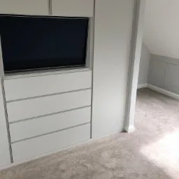 Biała zabudowa meblowa z wbudowanym telewizorem i szufladami w pokoju na poddaszu, jasna wykładzina dywanowa.