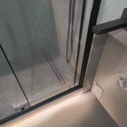Trowelmaster - Nowoczesna kabina prysznicowa z odpływem liniowym i deszczownicą, wykończona w szarościach. Widoczne detale: czarne ramy, minimalistyczny design, oświetlenie podłogowe.