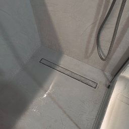 Trowelmaster - Szara kabina prysznicowa z odpływem liniowym i śladami wody, wykończona w stylu imitującym marmur. Widoczny wąż prysznicowy i fragment szklanej ścianki.