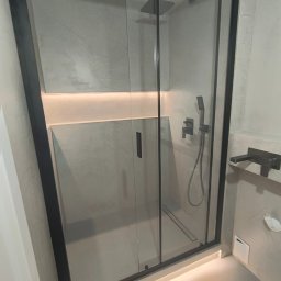 Trowelmaster - Nowoczesna kabina prysznicowa z czarnymi profilami i deszczownicą, wykończona szarym mikrocementem, z subtelnym oświetleniem LED u dołu i wnęce. Minimalistyczny design.