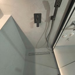 Trowelmaster - Nowoczesna kabina prysznicowa z czarną armaturą i odpływem liniowym, wykończona w jasnych odcieniach szarości z efektem dekoracyjnym na ścianach.