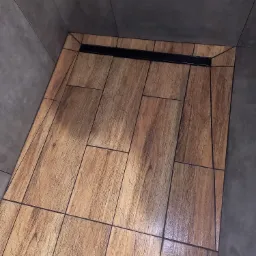 Wykładzina prysznicowa z płytek imitujących drewno, z czarnym odpływem liniowym, ułożona na szaro-betonowych ścianach łazienki.