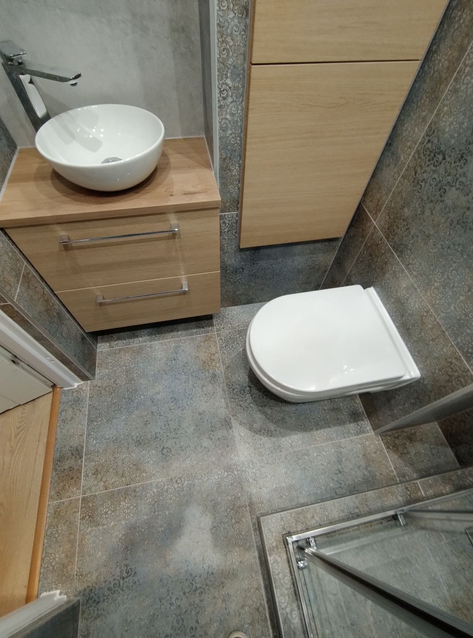 Nowoczesna łazienka z umywalką nablatową, szafką z drewnianym frontem, wiszącą toaletą i szklaną kabiną prysznicową, wykończona szaro-beżowymi płytkami z delikatnym wzorem.