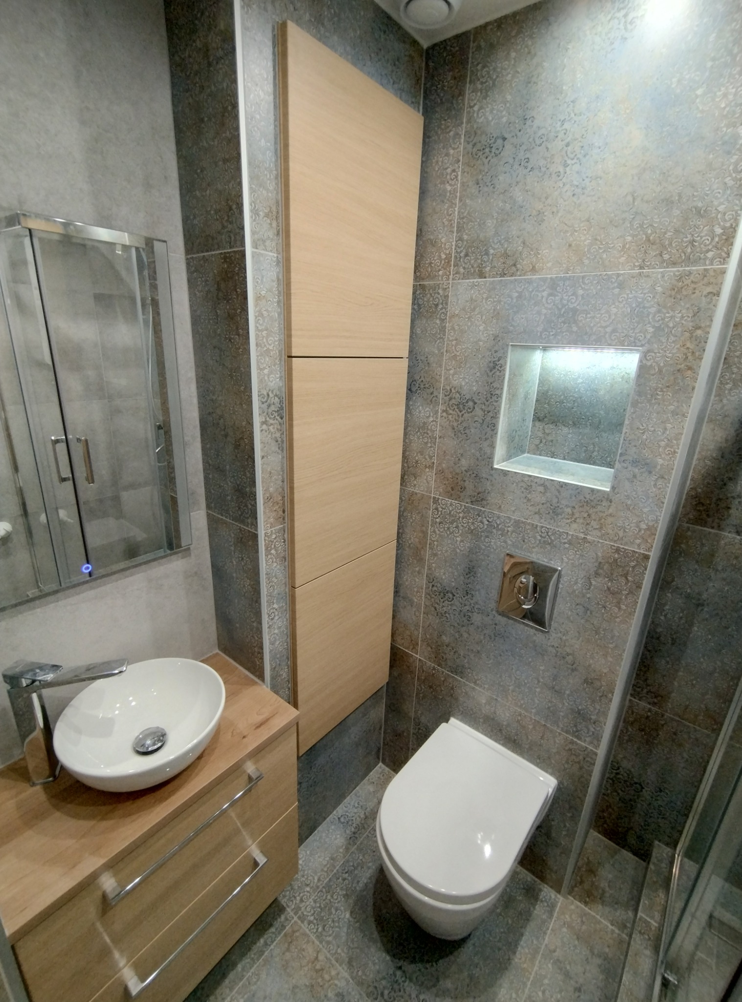 Nowoczesna łazienka z wiszącą toaletą, umywalką nablatową na szafce z drewnopodobnym fornirem, kabiną prysznicową ze szklanymi drzwiami oraz wysoką szafką w zabudowie, wykończona płytkami...