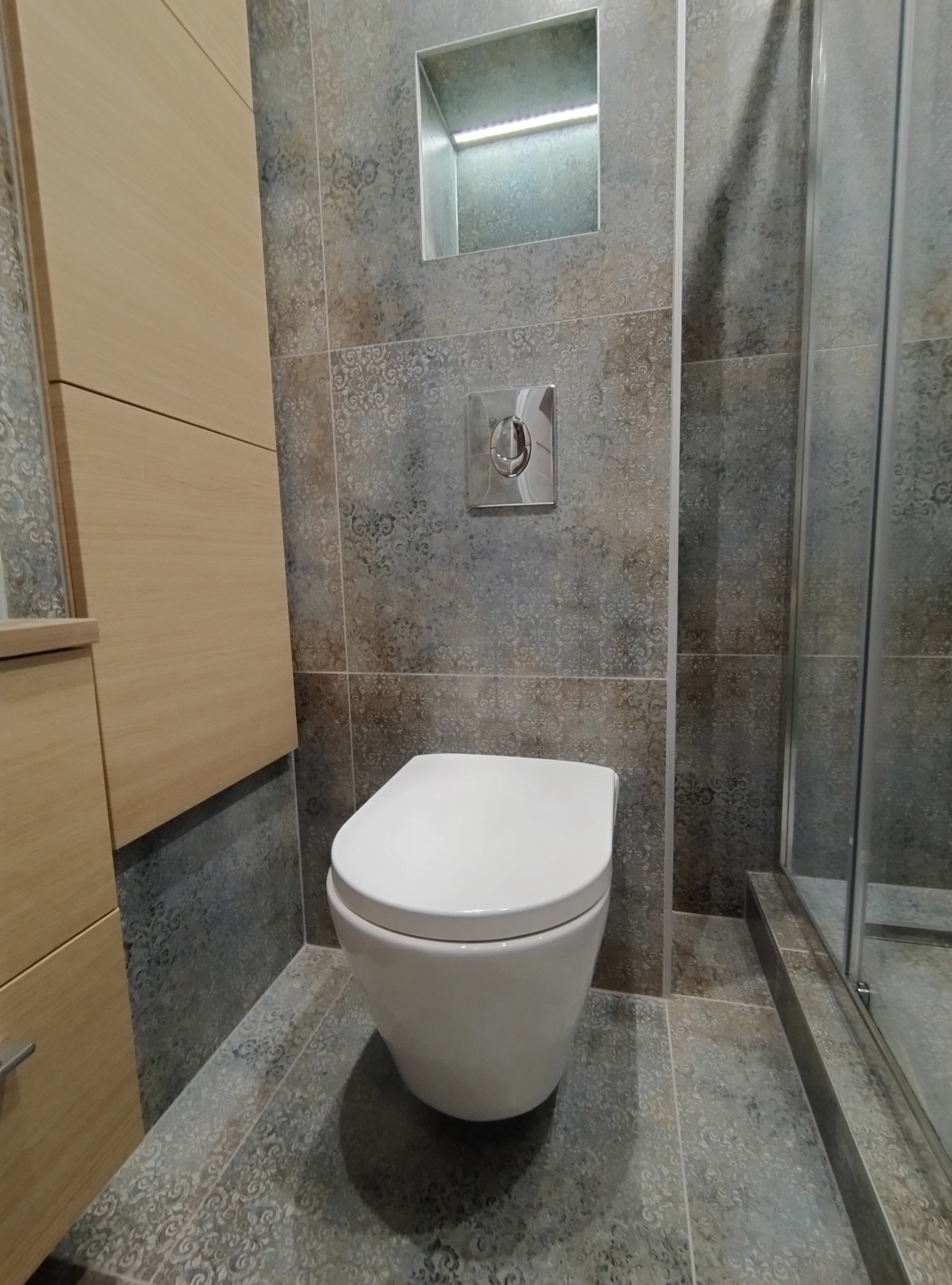 Wisząca toaleta w łazience wykończonej płytkami w odcieniach szarości i beżu, z zabudowaną spłuczką i wnęką z oświetleniem LED, obok kabina prysznicowa z metalowym profilem.