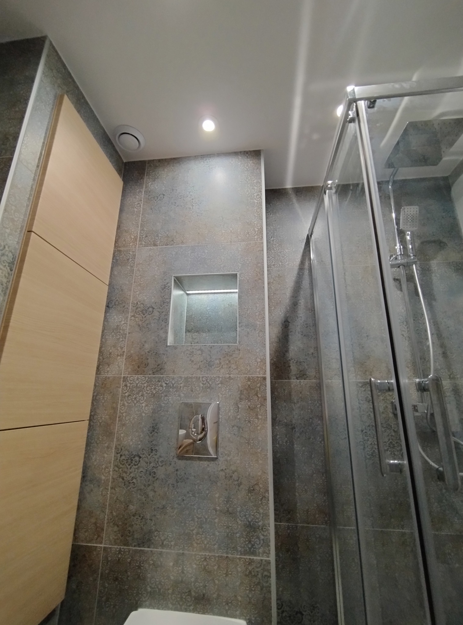 Wykończona łazienka z kabiną prysznicową, zabudową meblową w odcieniach beżu i szarymi płytkami z delikatnym wzorem, wnęka w ścianie z oświetleniem.