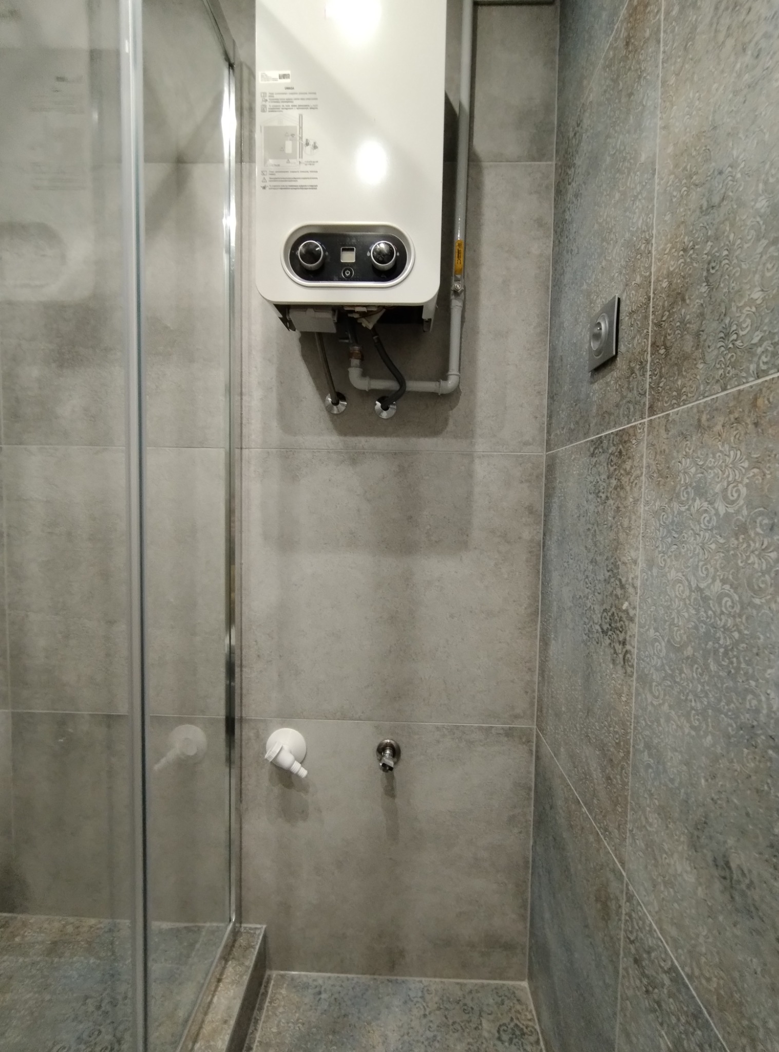 Szara łazienka z kabiną prysznicową, płytkami o wzorze ornamentowym na jednej ścianie, biały piec gazowy z czarnymi pokrętłami zamontowany na ścianie.