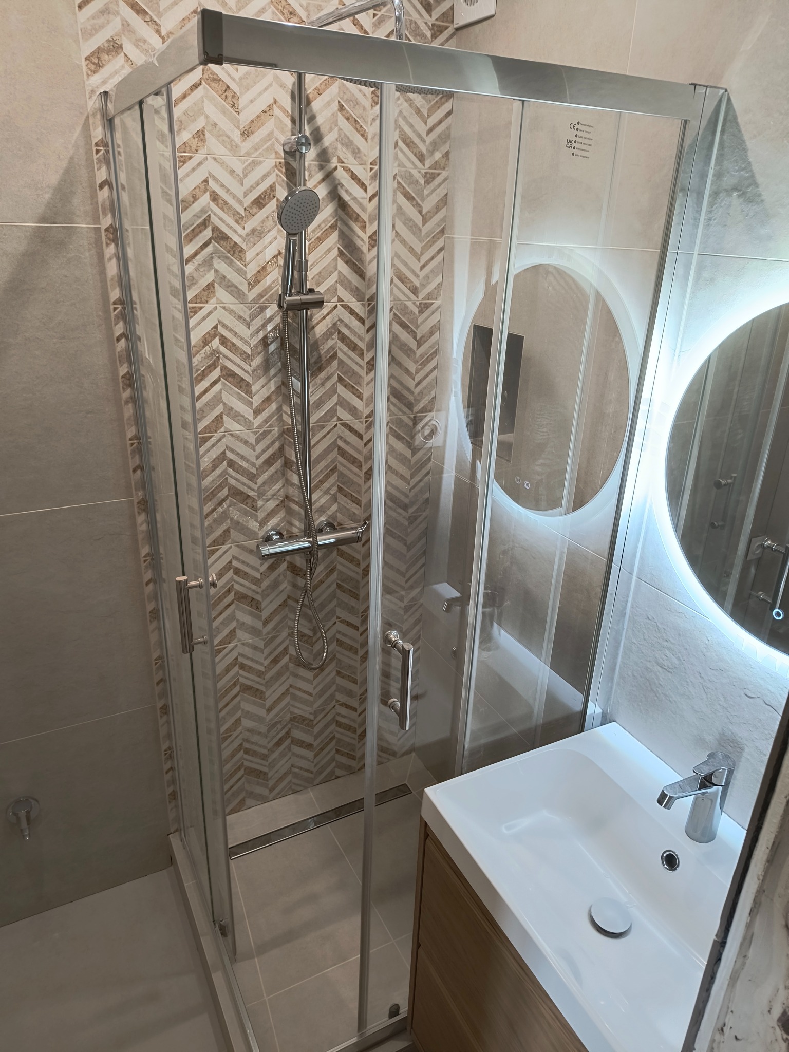 Nowoczesna kabina prysznicowa z chromowaną armaturą, wzorzyste płytki w jodełkę, okrągłe lustro z podświetleniem LED i biała umywalka nablatowa z drewnianą szafką w wykończonej łazience.