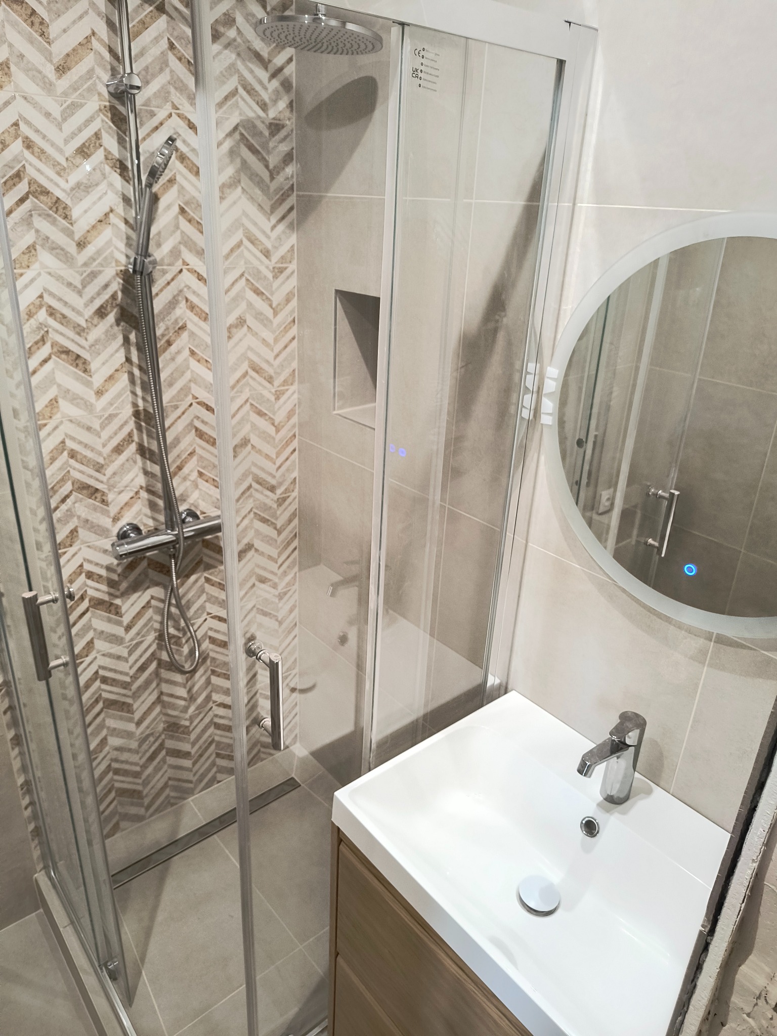 Nowoczesna łazienka z kabiną prysznicową z deszczownicą, umywalką nablatową i okrągłym lustrem z podświetleniem LED; ściana prysznica wykończona płytkami w jodełkę.