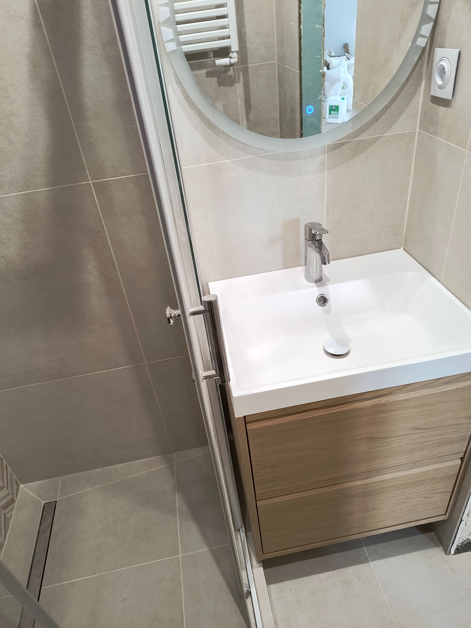 Nowoczesna łazienka z beżowymi płytkami, kabiną prysznicową ze szklanymi drzwiami, okrągłym lustrem z podświetleniem i umywalką z szafką z jasnego drewna.