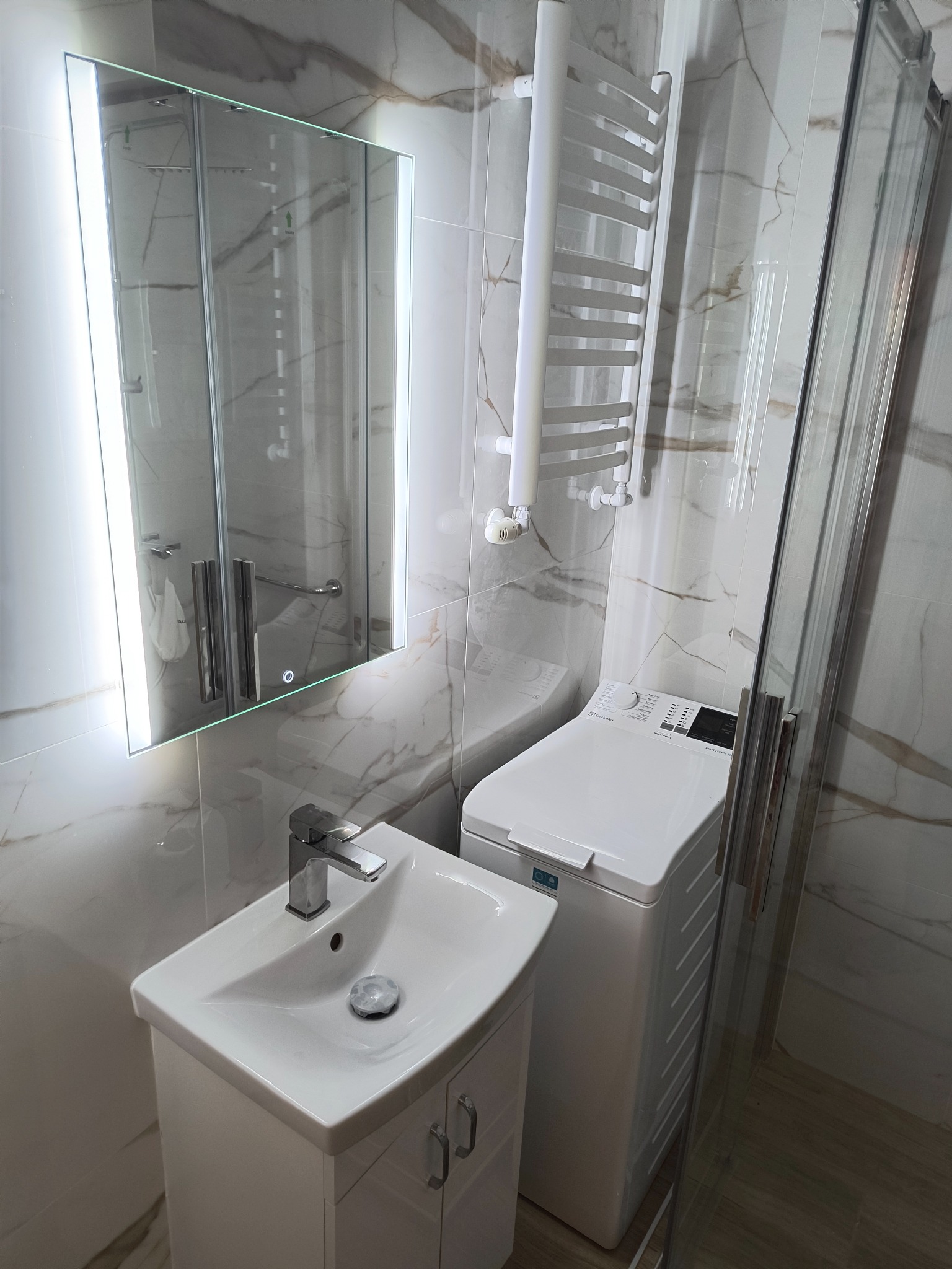 Biała umywalka z szafką, pralka i grzejnik drabinkowy w nowoczesnej łazience z marmurowymi płytkami i podświetlanym lustrem.