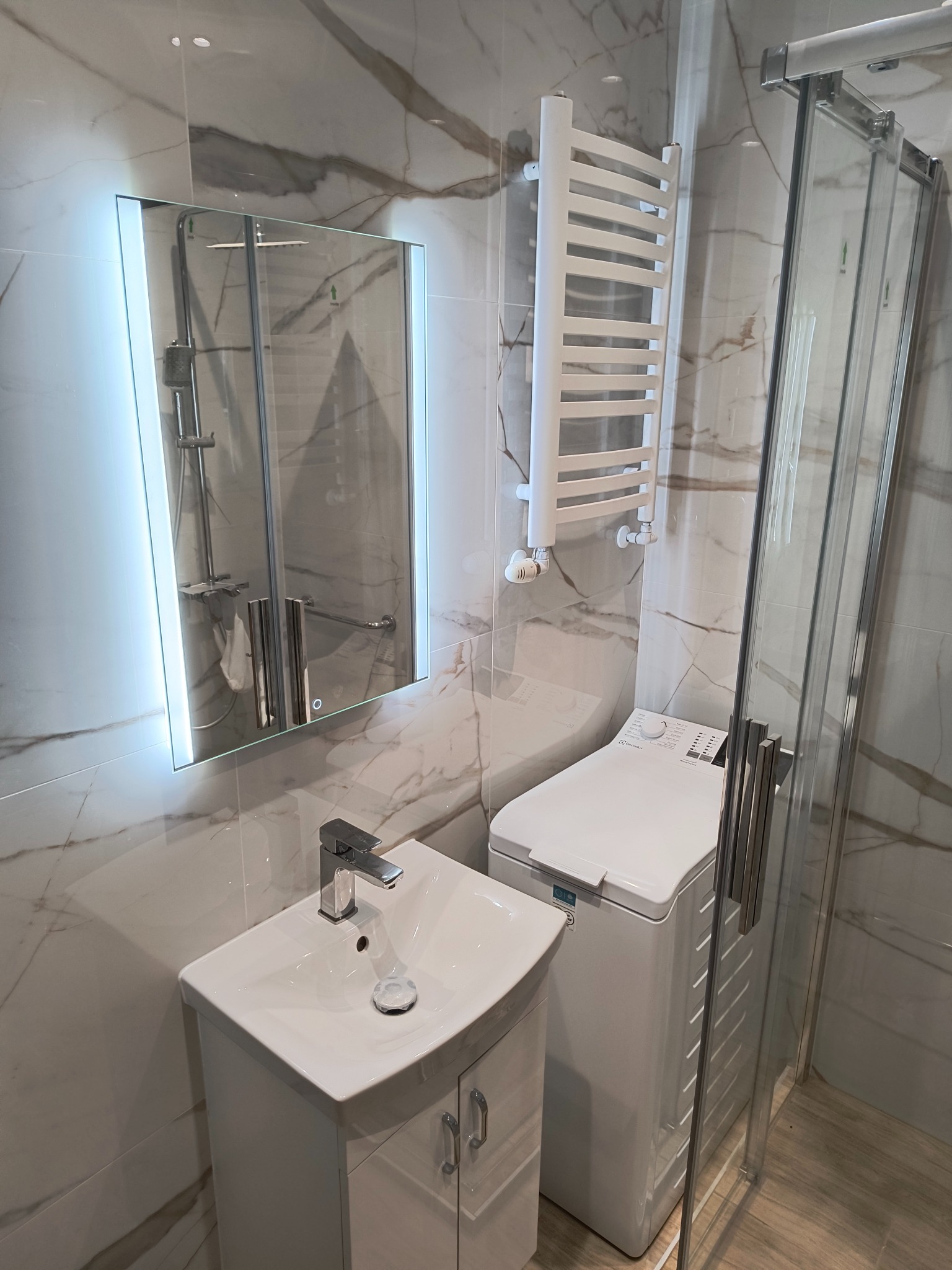 Nowoczesna łazienka z marmurowymi płytkami, podświetlanym lustrem, białą umywalką, grzejnikiem drabinkowym i pralką pod prysznicem.