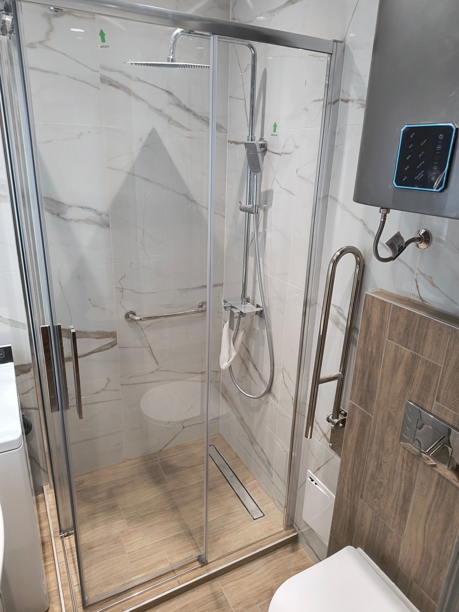 Nowoczesna kabina prysznicowa z marmurowymi płytkami, chromowanymi detalami i uchwytem dla niepełnosprawnych. Widoczny podgrzewacz wody z panelem dotykowym.