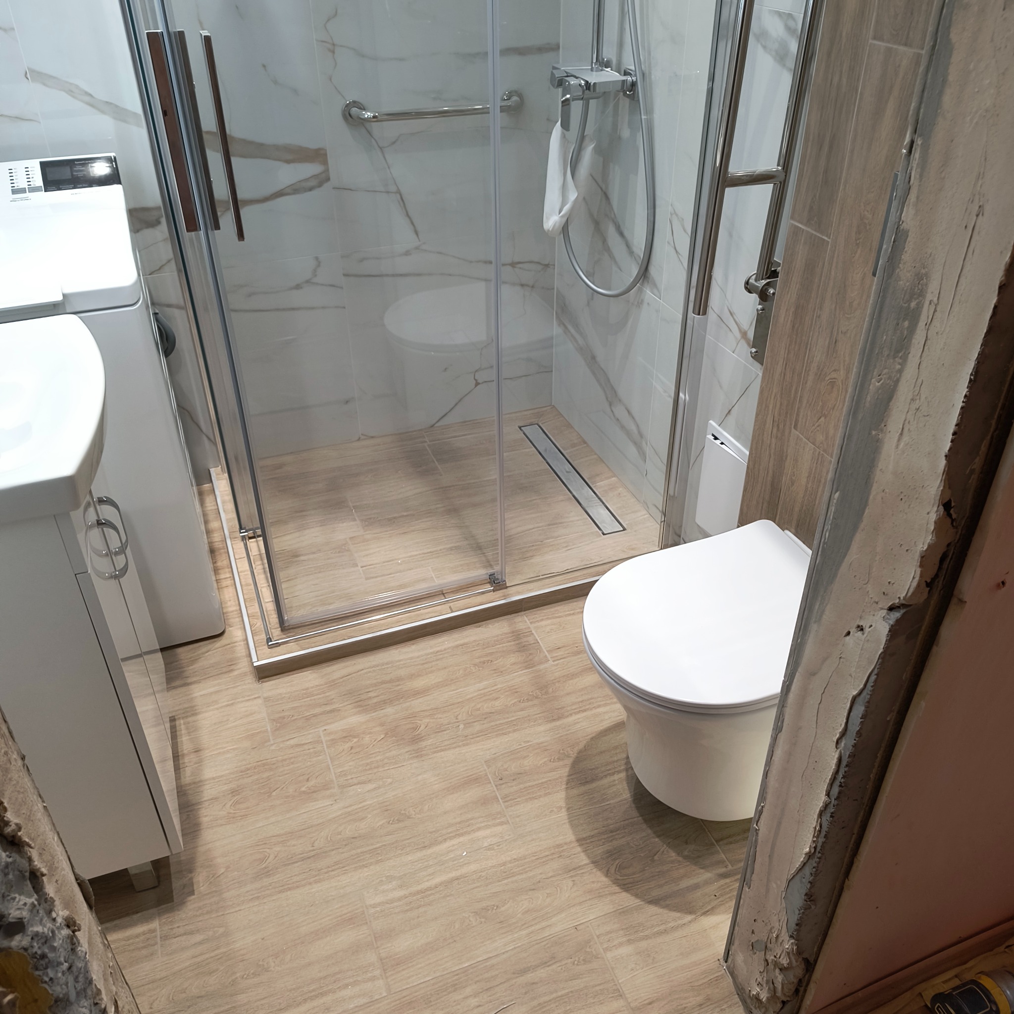 Wykończona łazienka z kabiną prysznicową, umywalką i toaletą. Uwagę zwracają płytki imitujące drewno na podłodze i marmur na ścianach. Widoczny fragment niedokończonego wykończenia.